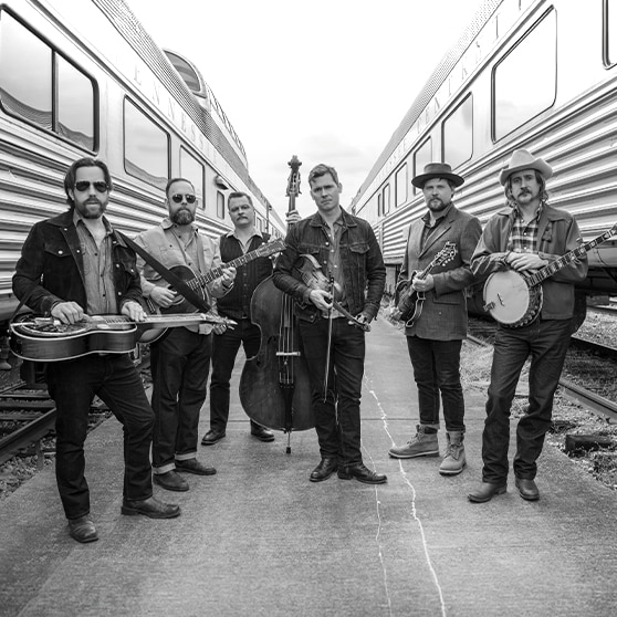 Six men standing in between two tour busses with instruments like banjos, guitars, and a double bass.