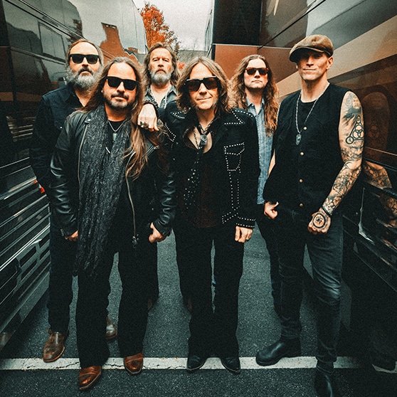 A group of seven adults stand outdoors in a narrow lane between two large tour buses. All wear dark, layered outfits, several with sunglasses, one with a hat, and one with visible arm tattoos. The group faces the camera, arranged in a loose line, with pavement below and trees and bus windows visible in the background.