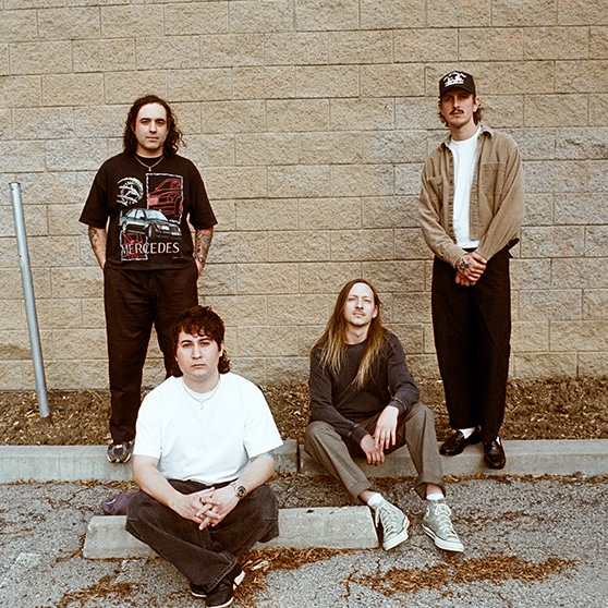 Four people are positioned in front of a tan brick wall in an outdoor parking lot. Two are standing—one wearing a black Mercedes graphic t-shirt and the other in a light jacket and white shirt. The other two are seated on the curb, one in a white t-shirt and dark pants, and the other in a long-sleeve shirt and sneakers.